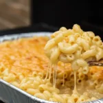 A wooden spoon lifting a large, cheesy scoop of smoked macaroni and cheese from an aluminum pan on a smoker grill, showing a perfect cheese pull.